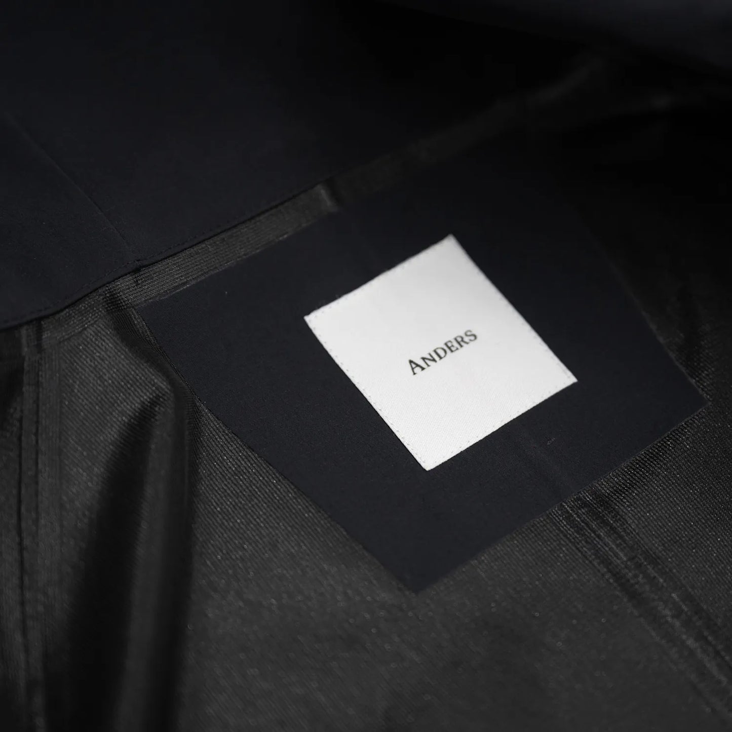 米軍の「ECWCS LEVEL6 JACKET GEN1 後期型」をベースにAndersが再構築したの3LAYERコート「3LAYERS ECWCS HARD SHELL COAT」のMATT BLACKカラーの襟裏ブランドロゴの写真
