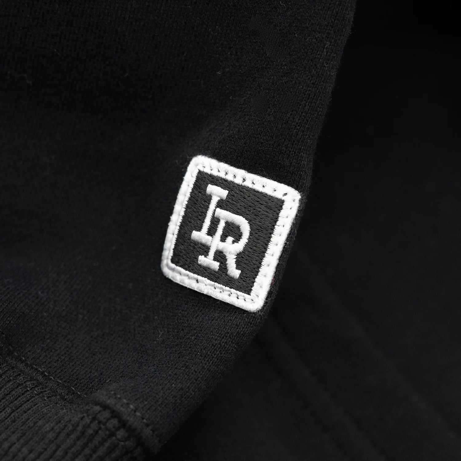 Liberaiders(リベレイダース)のフロントにデザインがプリントされた肉厚なフーディ(パーカー)「LR EMBROIDERY HOODIE」のBLACKカラーの左袖口のRロゴの写真